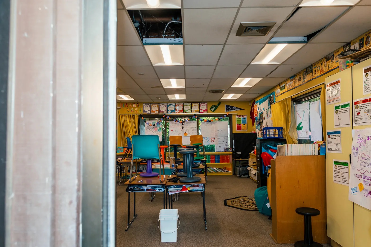 A view inside a classroom with a white bucket on the floor below a missing ceiling panel. 