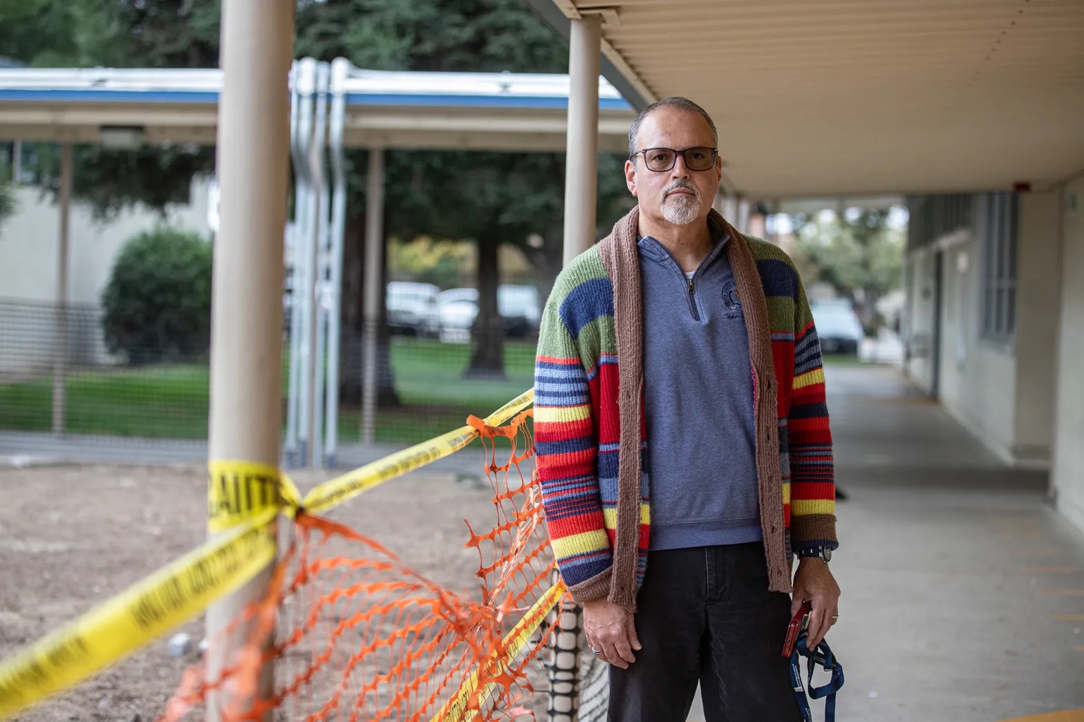 A middle-aged Latino man in a multicolored sweater stands next to caution tape in a school courtyard.